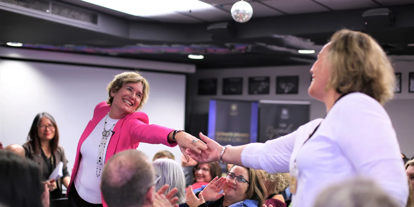 Remi reaching across a crowded room to shake a participant’s hand